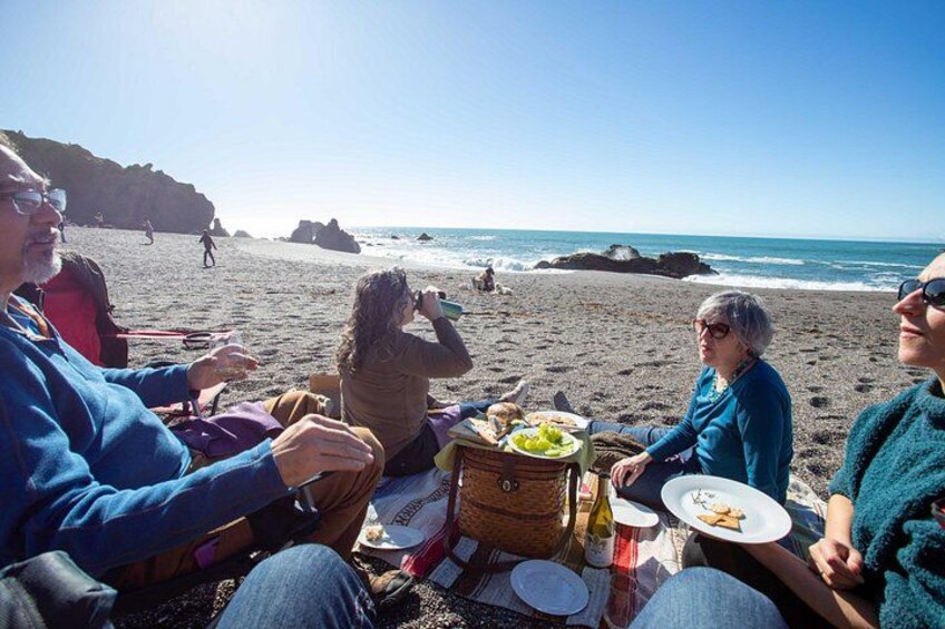 A group of people sitting on a beach with a picnic setup, including plates, a basket, and a bottle. 4