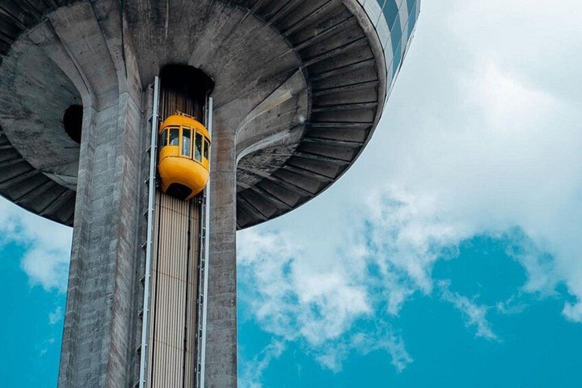 Un teleférico amarillo asciende por una alta torre de hormigón bajo un cielo azul con nubes. 1