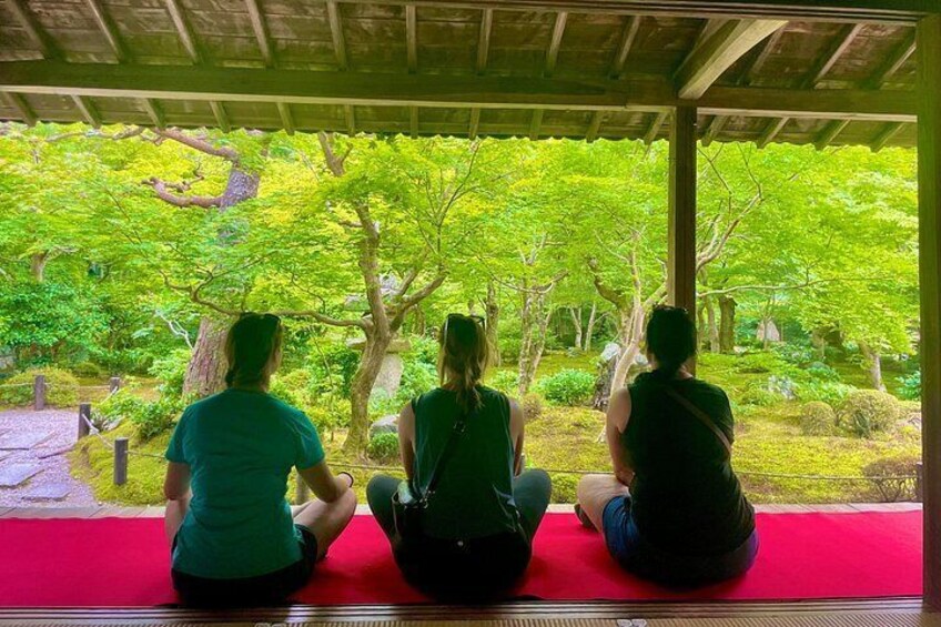 Three people sitting on a red mat inside a covered area, looking at a lush green garden. 4