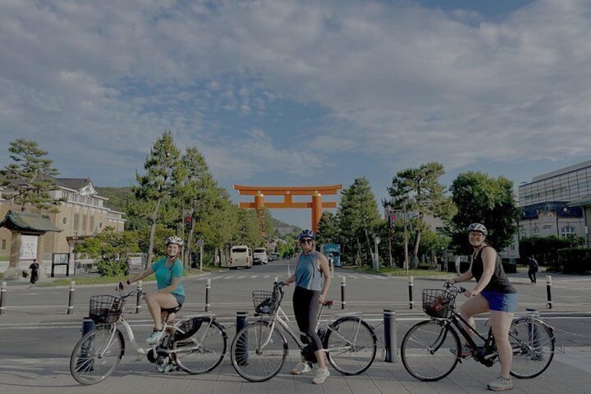 Three people on bicycles, a traditional Japanese torii gate, and a modern building in the background. 2