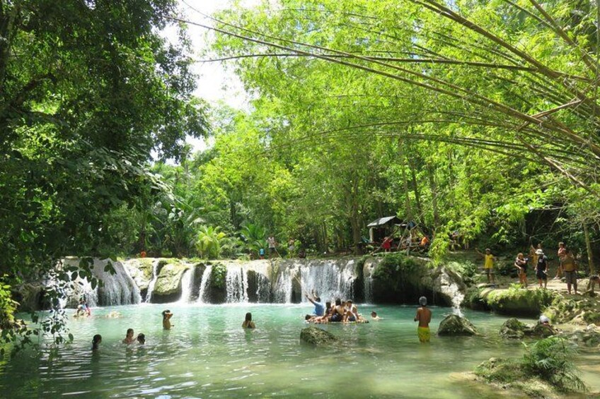A waterfall, clear water, and people swimming and relaxing in a forested area. 2