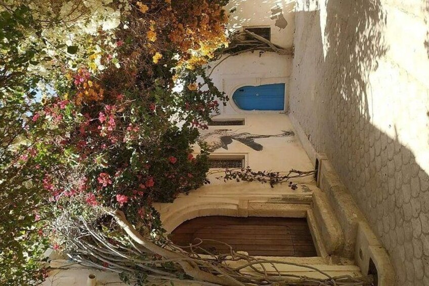 A narrow alleyway with a blue door, a wooden gate, and a tree with pink flowers. 2