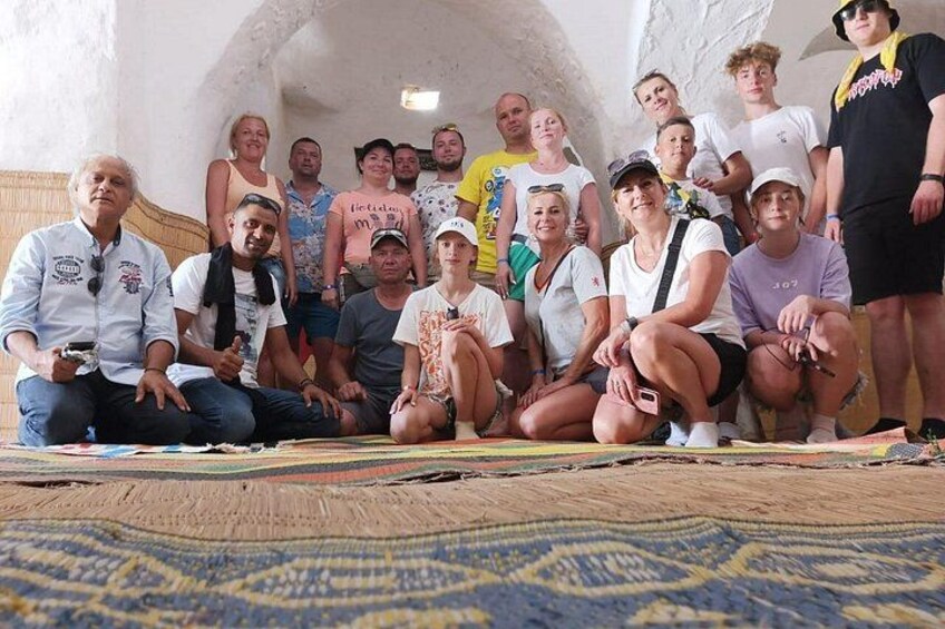 Un groupe de personnes posant pour une photo dans une pièce dont le sol est recouvert d’un tapis à motifs et les murs sont en pierre. 2