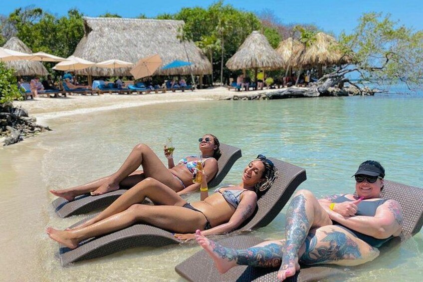 Tres personas tomando el sol en sillones de playa, con cabañas de techo de paja al fondo. 5
