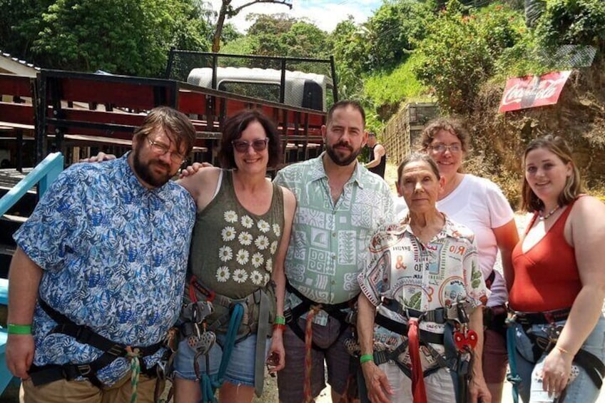 A group of people with harnesses, standing in an outdoor setting with trees and a structure in the background. 3