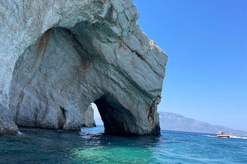 A large rock formation with a natural arch over the sea, clear blue water, and distant mountains. 5