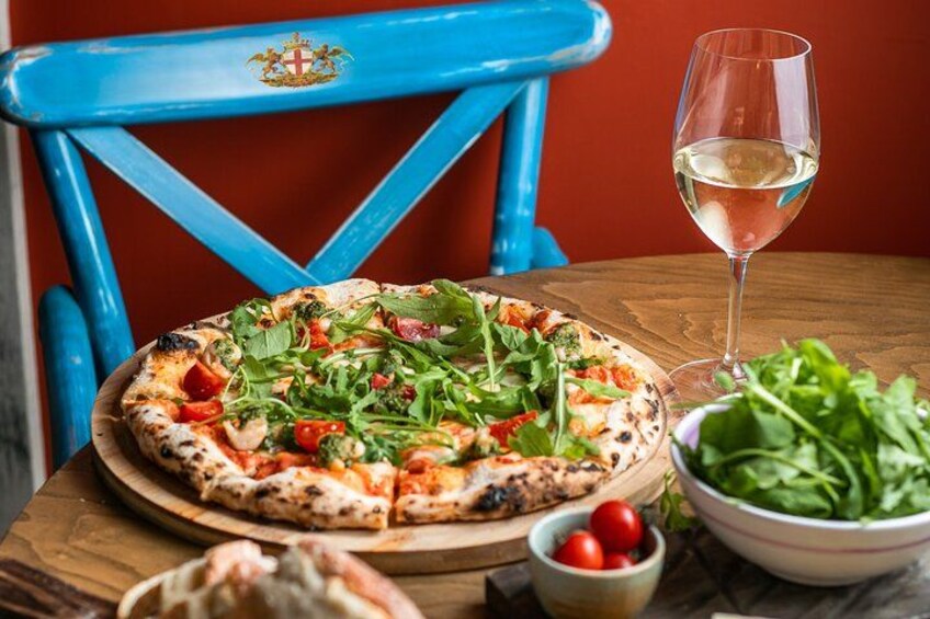 Una pizza sobre una tabla de madera, una copa de vino blanco y un recipiente con verduras sobre una mesa. 3