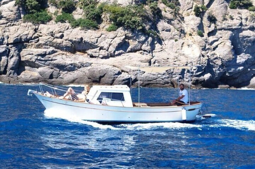A small white boat with two people on board, one at the front and one at the back, moving through a body of water with rocky cliffs in the background. 4