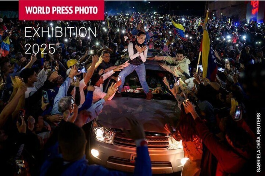 A crowd of people gathered around a car with a person on top. 2