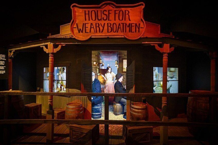 A wooden building with a sign that reads 'HOUSE FOR WEARY BOATMEN' and a bar area with stools and barrels. 5