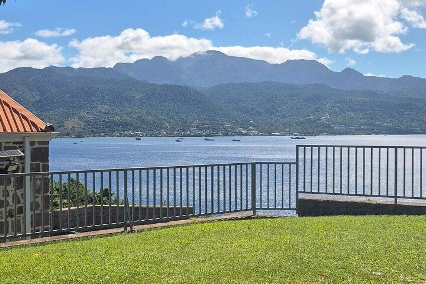 Ein grasbewachsenes Gelände mit einem Metallzaun, einem Steinhaus und Blick auf einen See mit Booten und Bergen im Hintergrund. 5