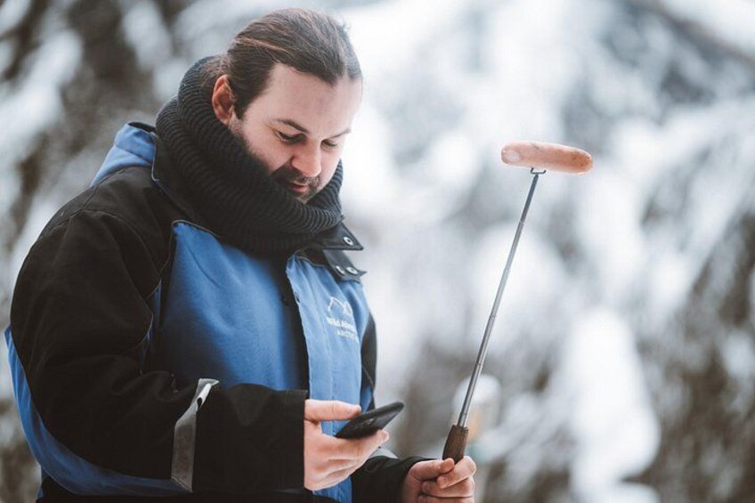 A person in a blue jacket holding a smartphone and a metal rod with a sausage on it. 2