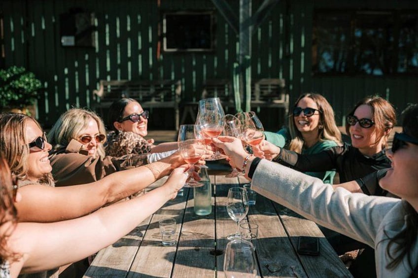 A group of people toasting with wine glasses around a wooden table outdoors. 2