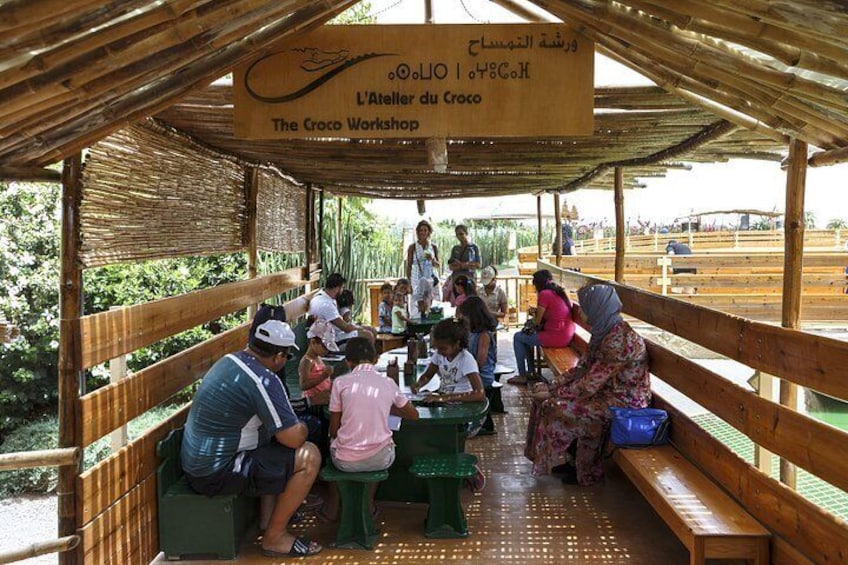A covered wooden structure with a sign reading 'The Croco Workshop.' People are seated at tables under the roof, and a few are standing. 4