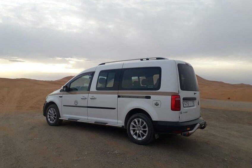 A white van parked on a desert road with sand dunes in the background. 1