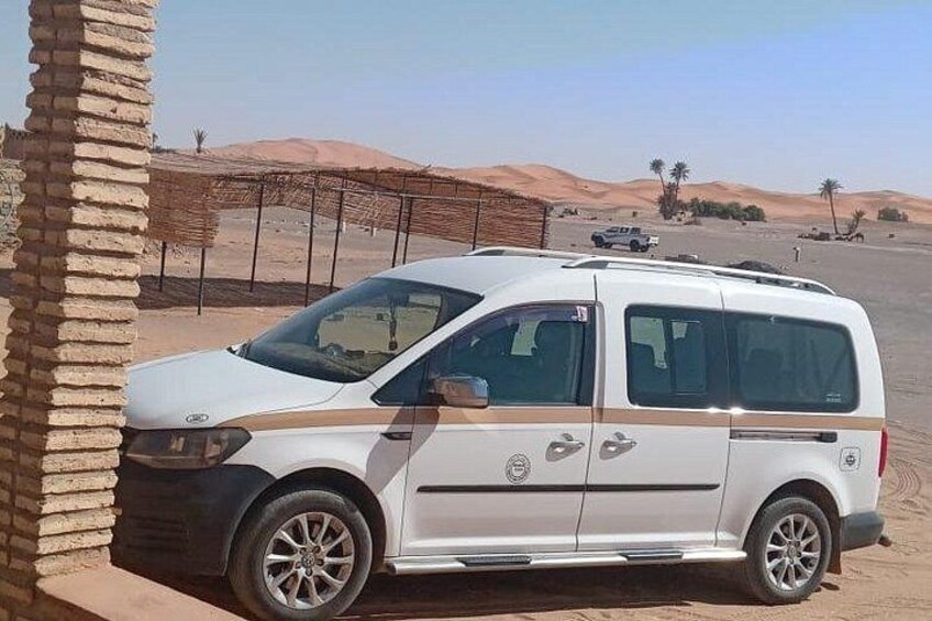 A white van parked in a desert with sand dunes and palm trees in the background. 2