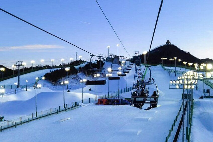 A ski lift with chairs moving up a snowy slope, illuminated lights, and a mountain in the background. 2