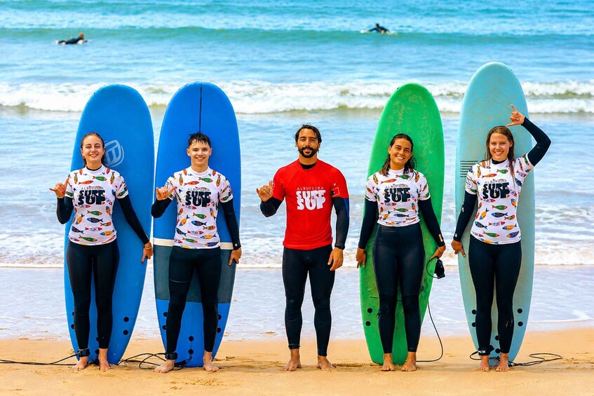 Five people on a beach holding surfboards, wearing matching shirts with 'ALQUIFERA SURF' printed on them. 1
