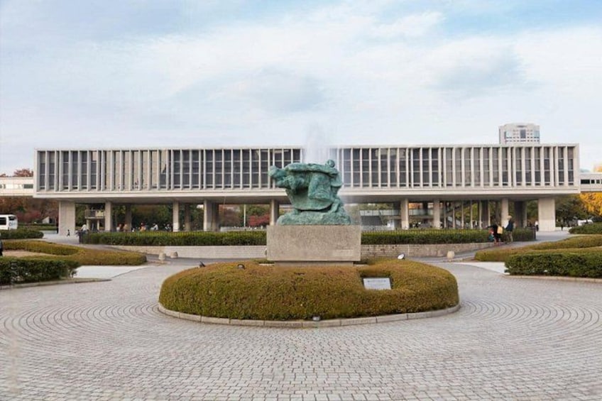 Een modern gebouw met een glazen gevel, een centraal geplaatst sculptuur en een rond aangelegde groenstrook. 3