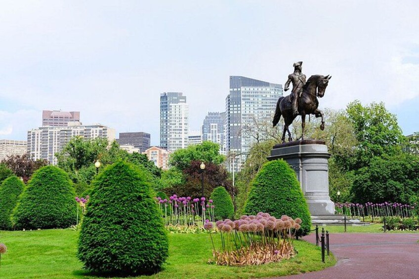 En statue af en person til hest i en park med velplejede buske og blomster, og en bysilhuet i baggrunden. 2
