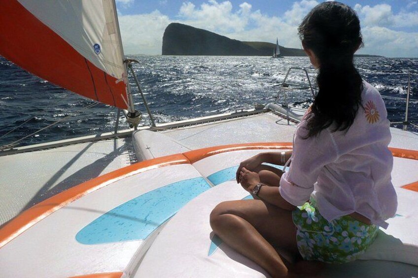 A person sitting on a boat with a sail, ocean, and a distant island. 4