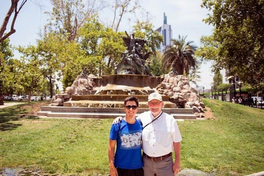 Dos personas de pie frente a una fuente con una estatua en un parque. 3
