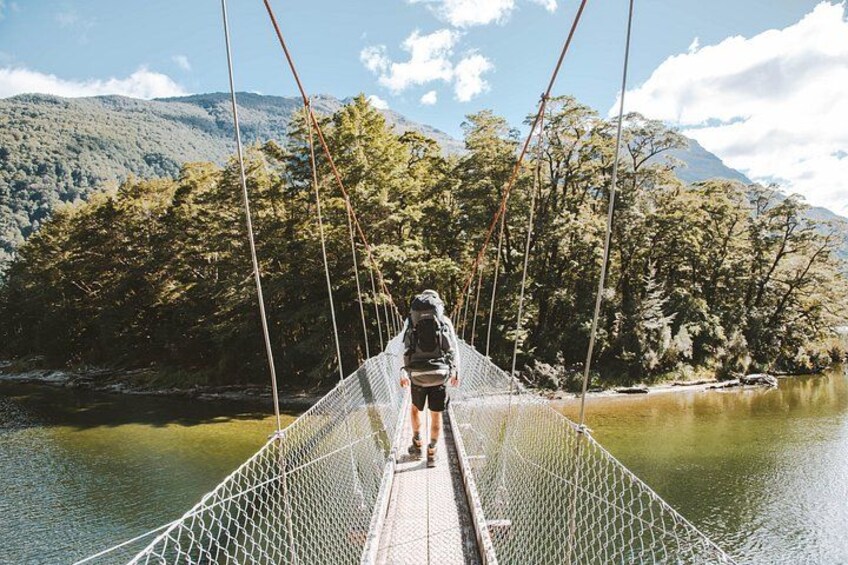 A person standing on a suspension bridge over a river, surrounded by trees and mountains. 5