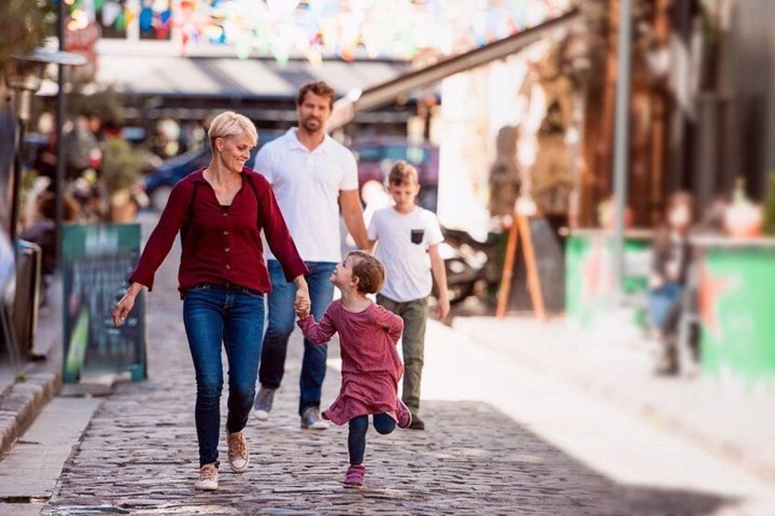 Eine Familie spaziert auf einem Kopfsteinpflasterweg, im Hintergrund Geschäfte und bunte Flaggen. 5