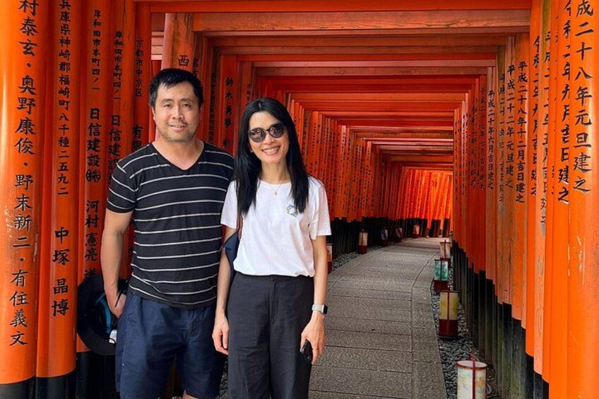 Two people standing in front of a row of orange torii gates with Japanese characters on them. 1