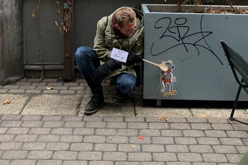 En person knæler ved siden af en væg med graffiti og et klistermærke med en tegneseriefigur. 3