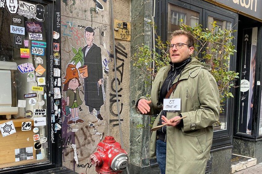 En person står foran en bygning med et muralmaleri, holder et skilt, og der er en rød brandhane i nærheden. 1