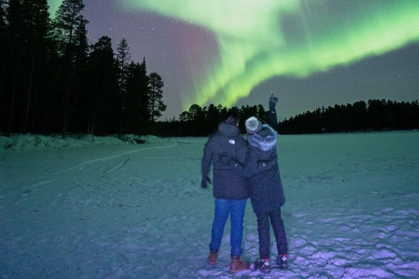 Two people standing on a snow-covered ground, looking at the night sky with the aurora visible above. 1