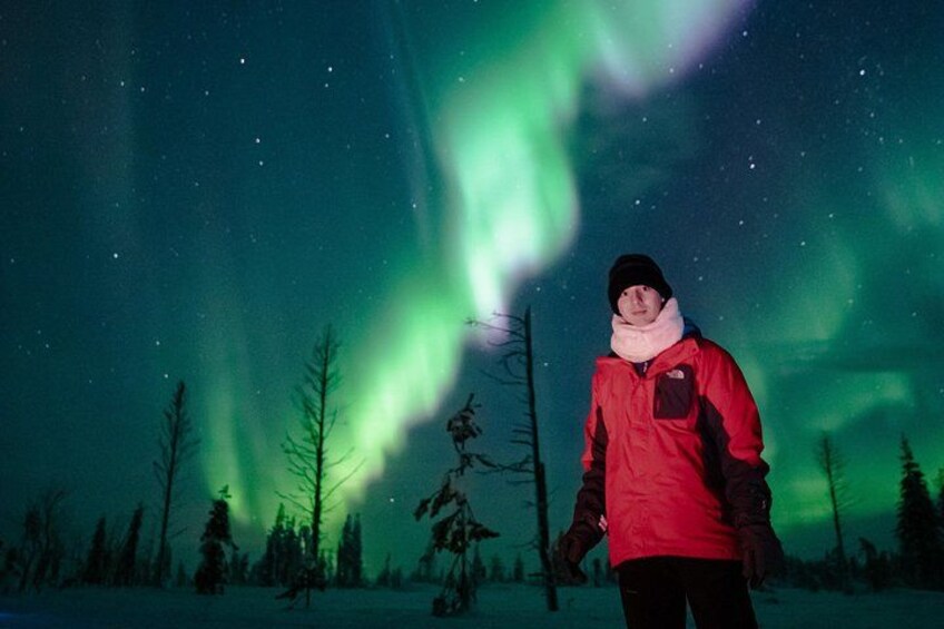 A person standing in a snowy landscape with trees and the Northern Lights in the background. 2