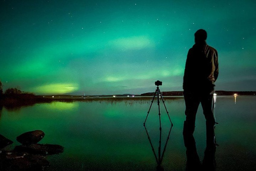 A person standing by a tripod with a camera, facing a body of water under a green aurora. 3