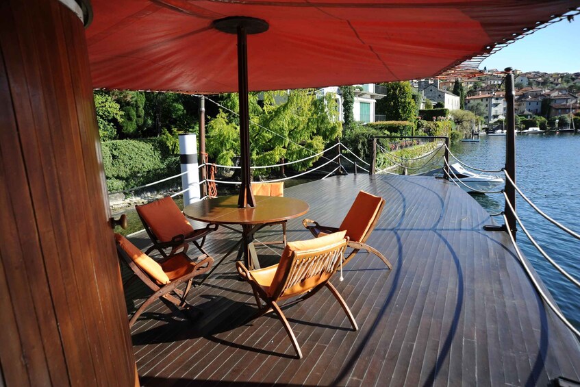 A wooden deck with a table and chairs under a red canopy, overlooking a river with buildings in the background. 2