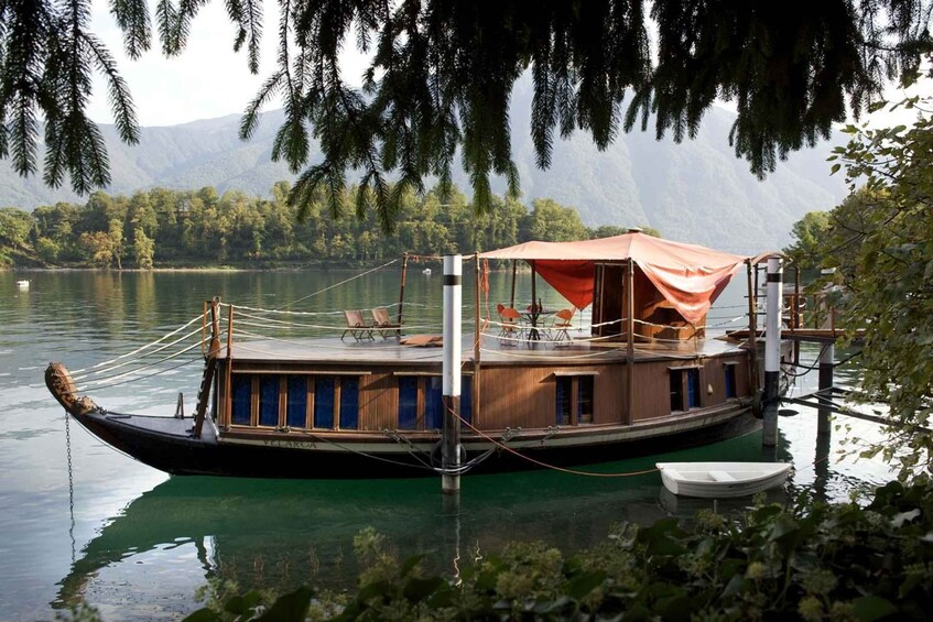 A wooden boat with a red canopy docked on a calm lake, surrounded by lush greenery and mountains in the background. 1