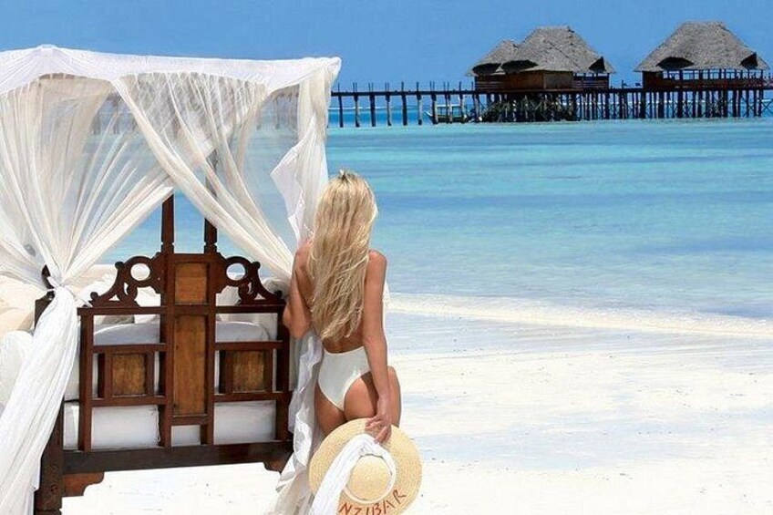 A beach scene with a bed, a straw hat, and overwater bungalows in the background. 3