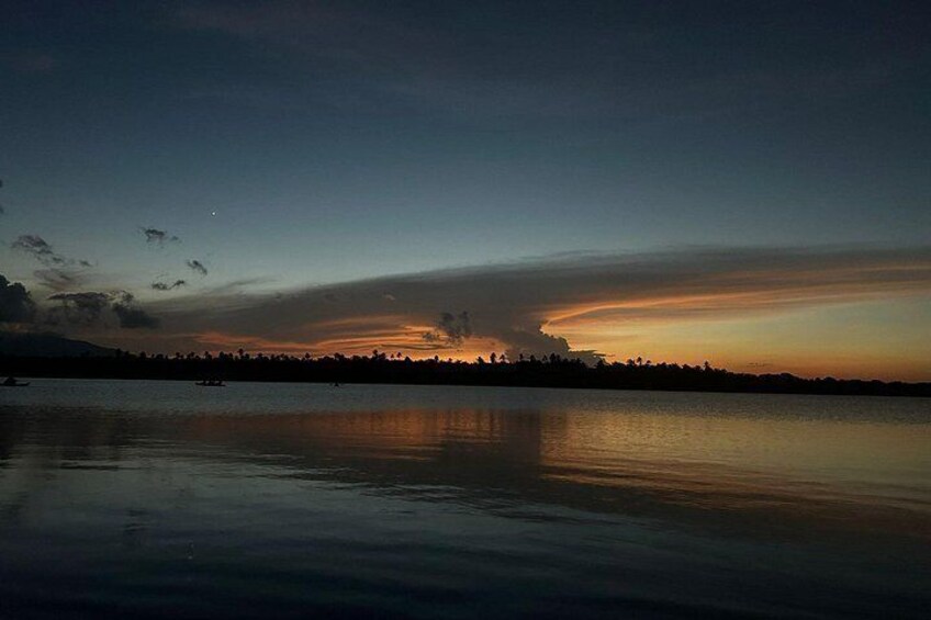 A calm lake at dusk with a sunset reflecting on the water, a silhouette of a distant shoreline, and a few boats on the water. 5