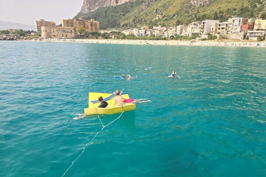 A yellow raft with people on a calm sea, a beach with buildings, and a mountainous backdrop. 3
