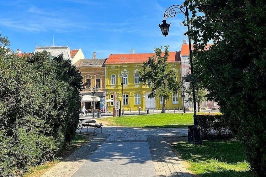 A paved pathway leading to a yellow building with a red roof, surrounded by green trees and a clear blue sky. 2