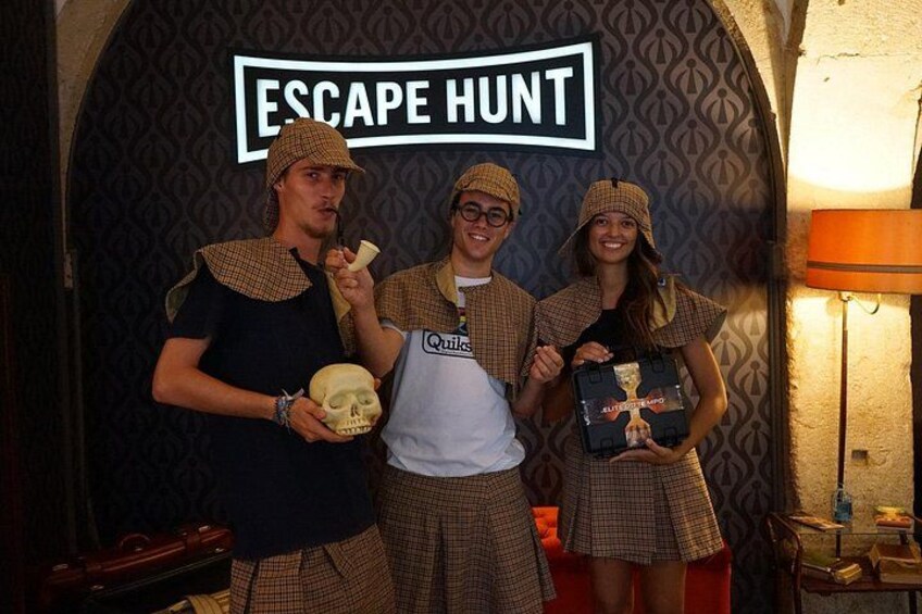 Trois personnes dans un décor thématique, avec une pancarte indiquant « ESCAPE HUNT ». 1