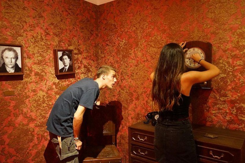 A man and a woman in a room with patterned wallpaper, a clock, and a dresser. 2