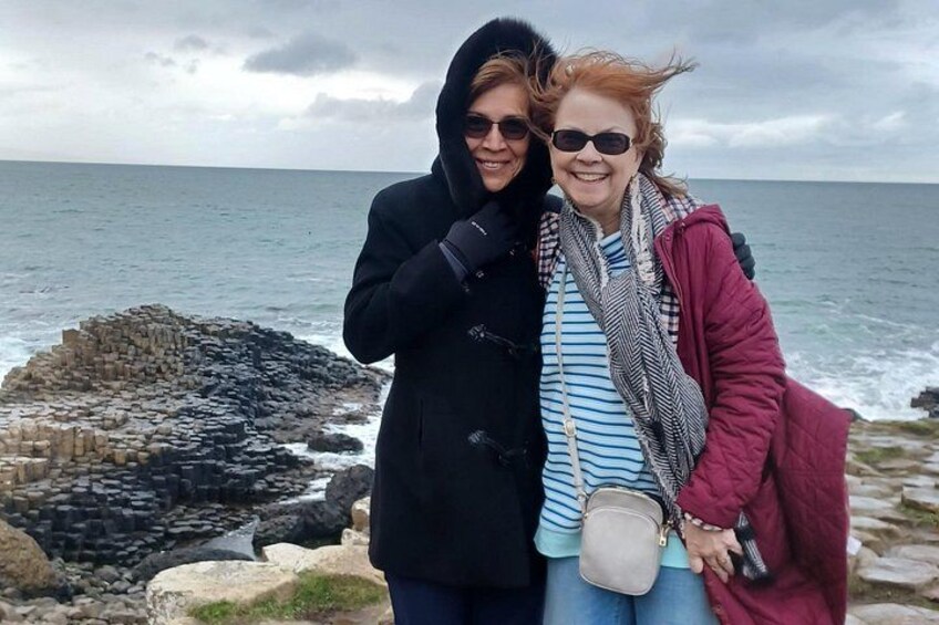 To personer står ved en stenfyldt kystlinje med havet i baggrunden. 2