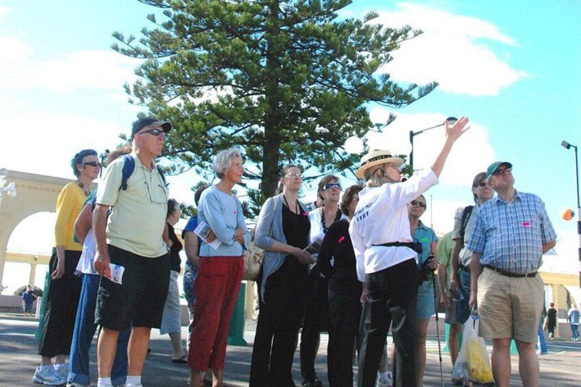 A group of people standing outdoors, some with backpacks, near a large tree and a bridge in the background. 3
