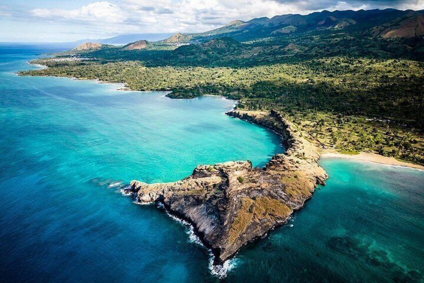 Eine Küstenlandschaft mit einem markanten Felsen, klarem türkisblauem Wasser und sanften grünen Hügeln im Hintergrund. 5