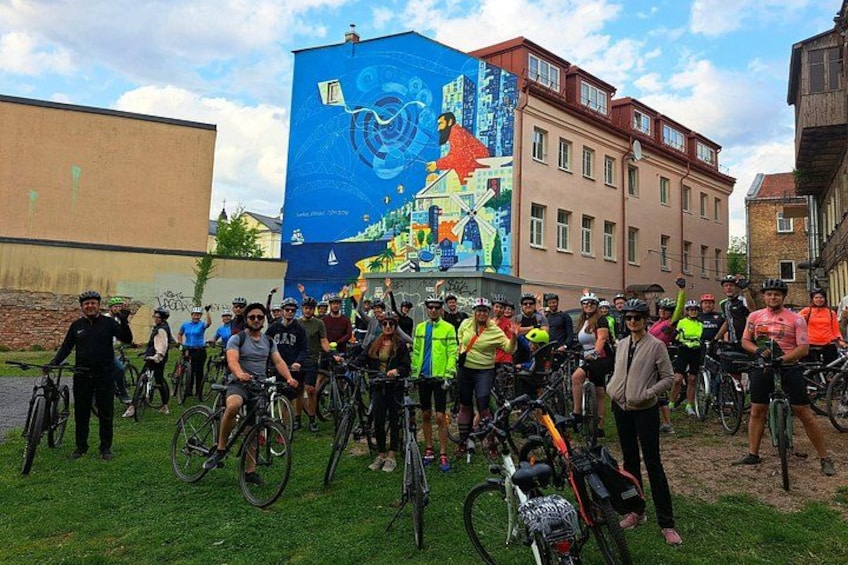 A group of cyclists gathered in an urban setting with a mural in the background. 3