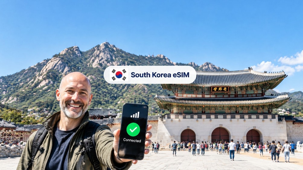 Un uomo tiene in mano uno smartphone con una spunta verde, in piedi di fronte a un edificio tradizionale coreano con montagne sullo sfondo. 1