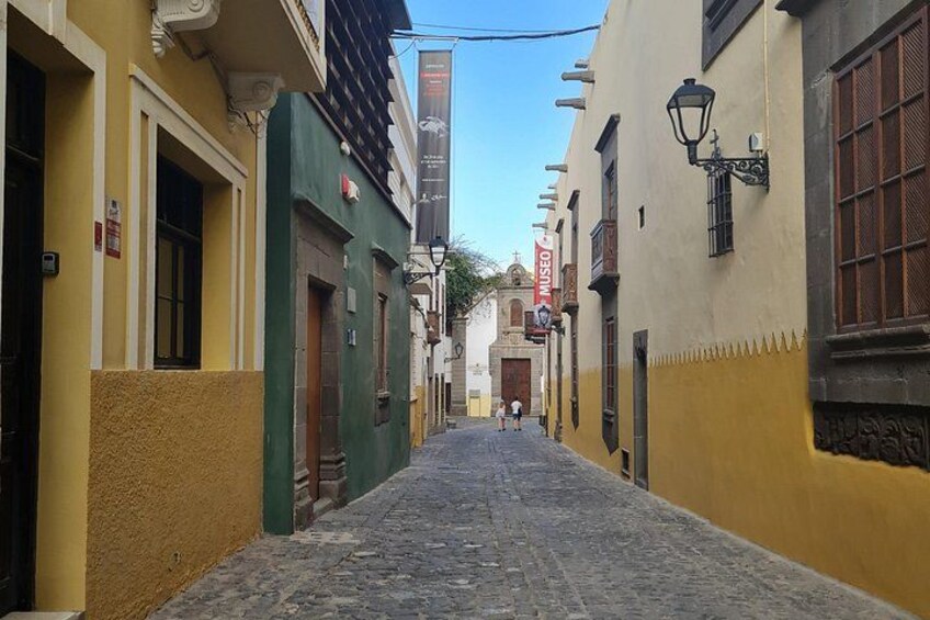 A narrow cobblestone street flanked by buildings with yellow and green facades, a church with a bell tower, and a red banner. 1