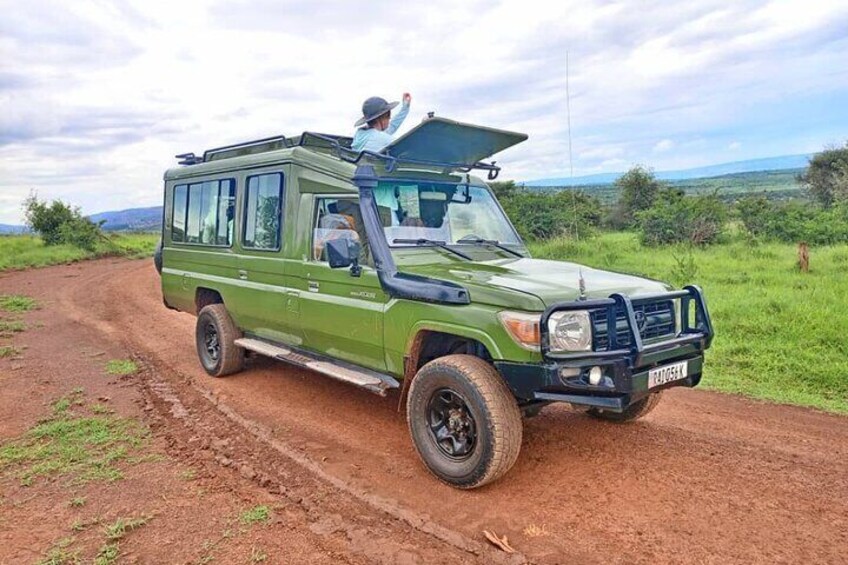 A green safari vehicle on a dirt road with a person standing on the roof. 3