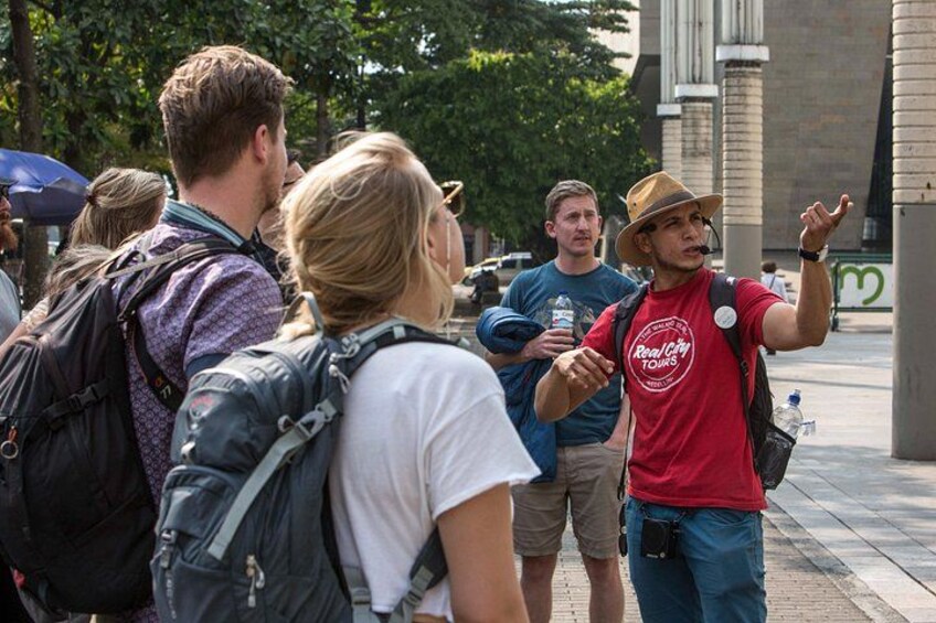 A group of people standing on a sidewalk, some with backpacks, one wearing a red shirt with a logo, and another in a hat. 1